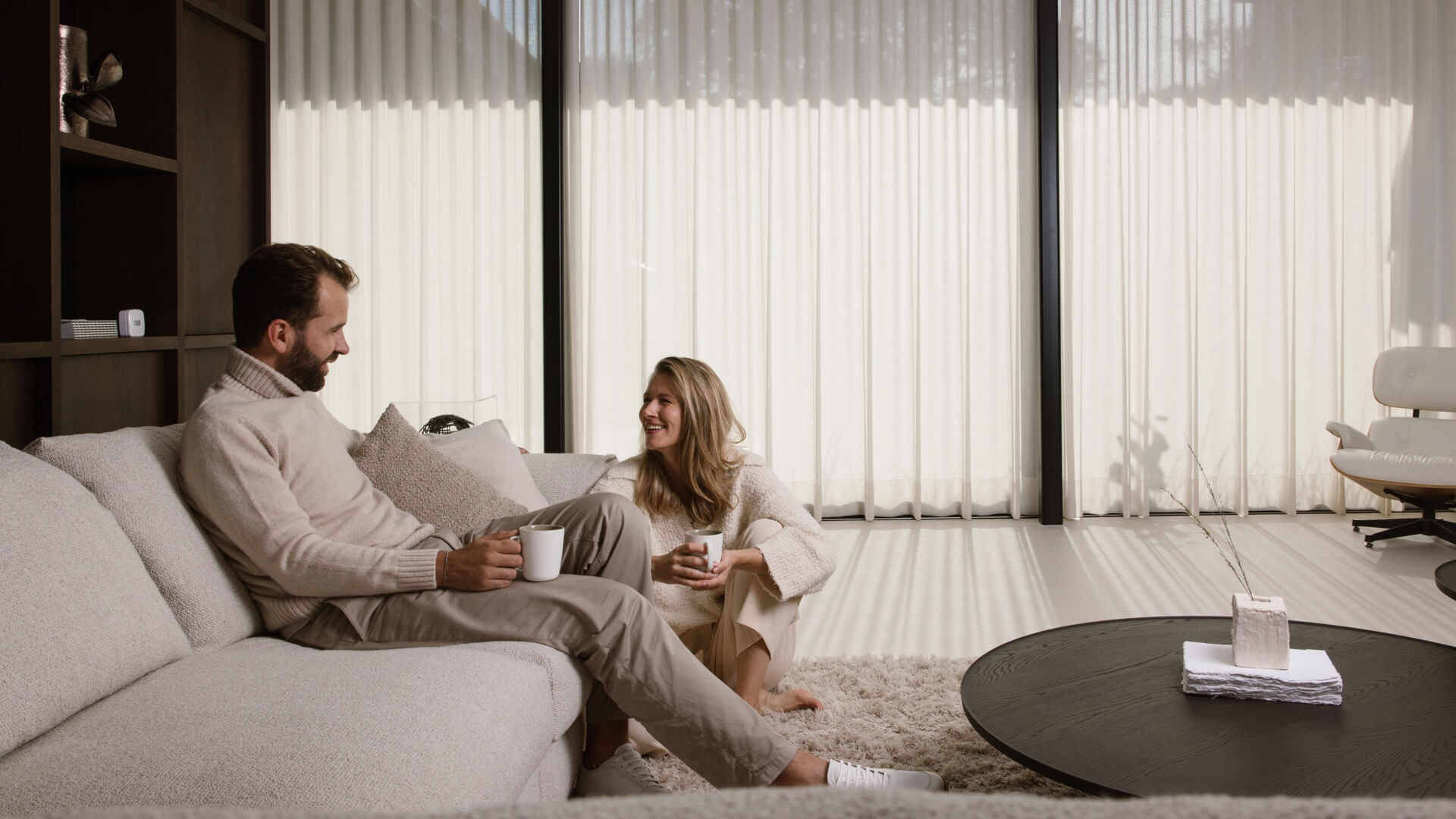 Two people sitting in a living room with floor-to-ceiling Motionblinds curtains filtering afternoon sunlight